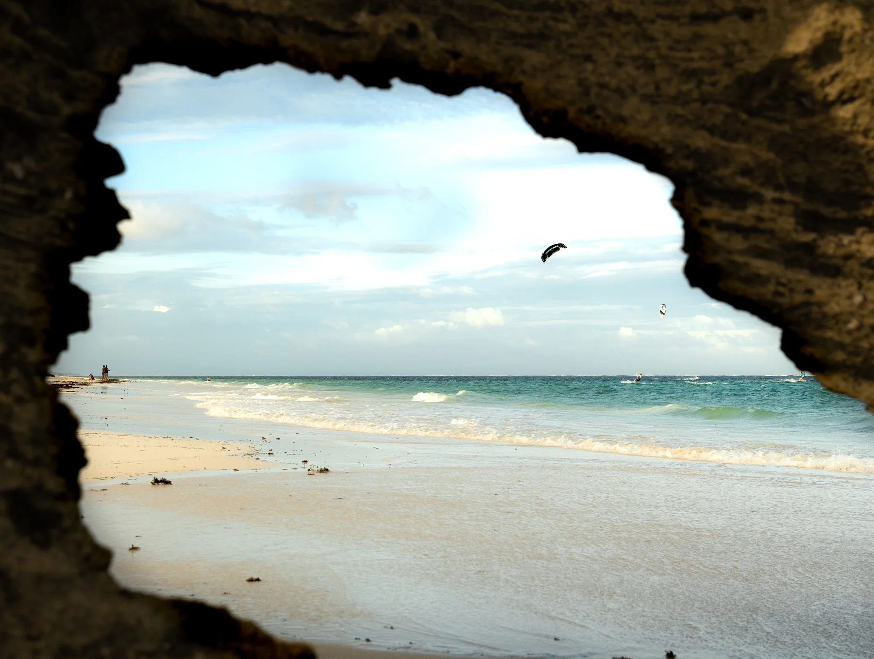 Blick durch eine Felsöffnung auf den weißen Strand und einen Kite